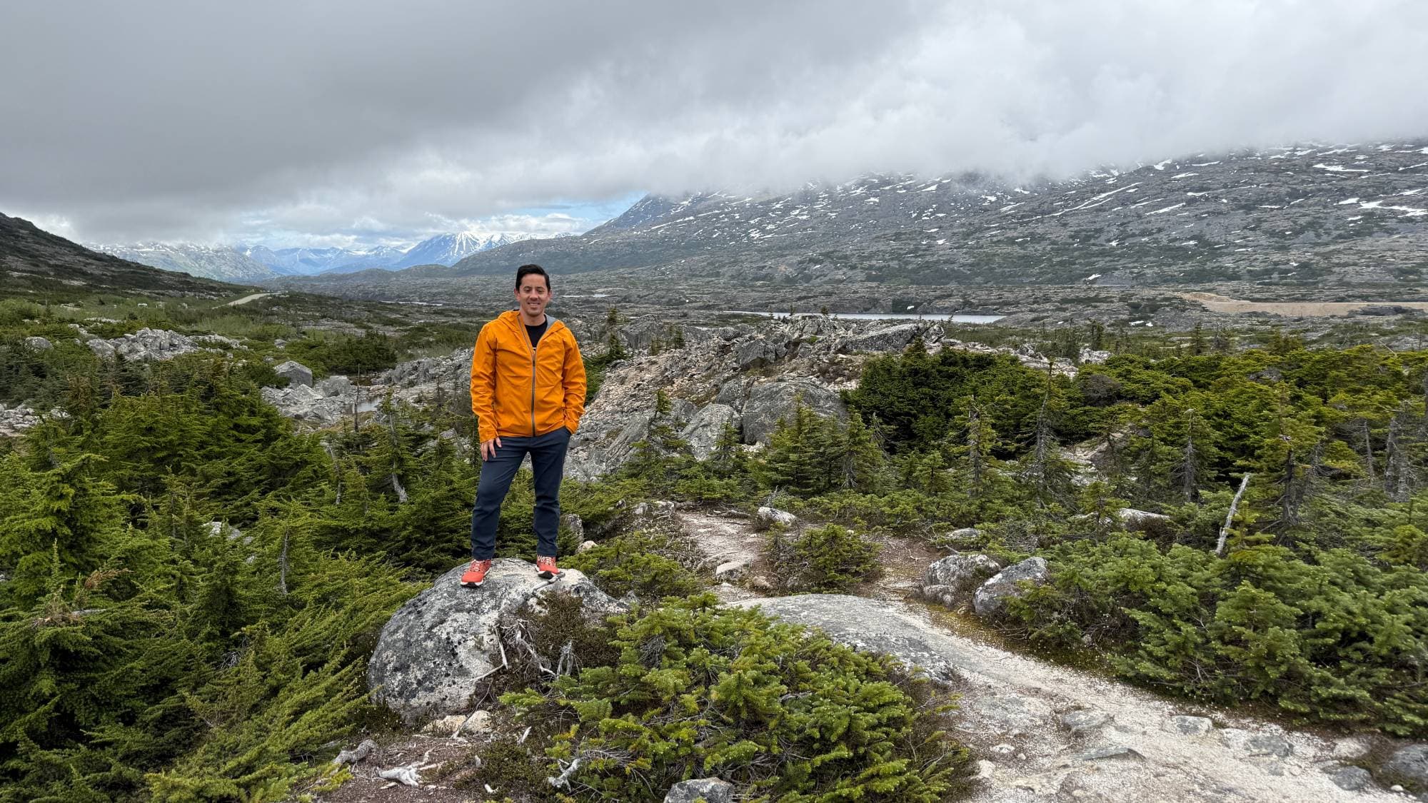 Kevin in an orange jacket standing on a boulder in a rocky alpine valley under low clouds