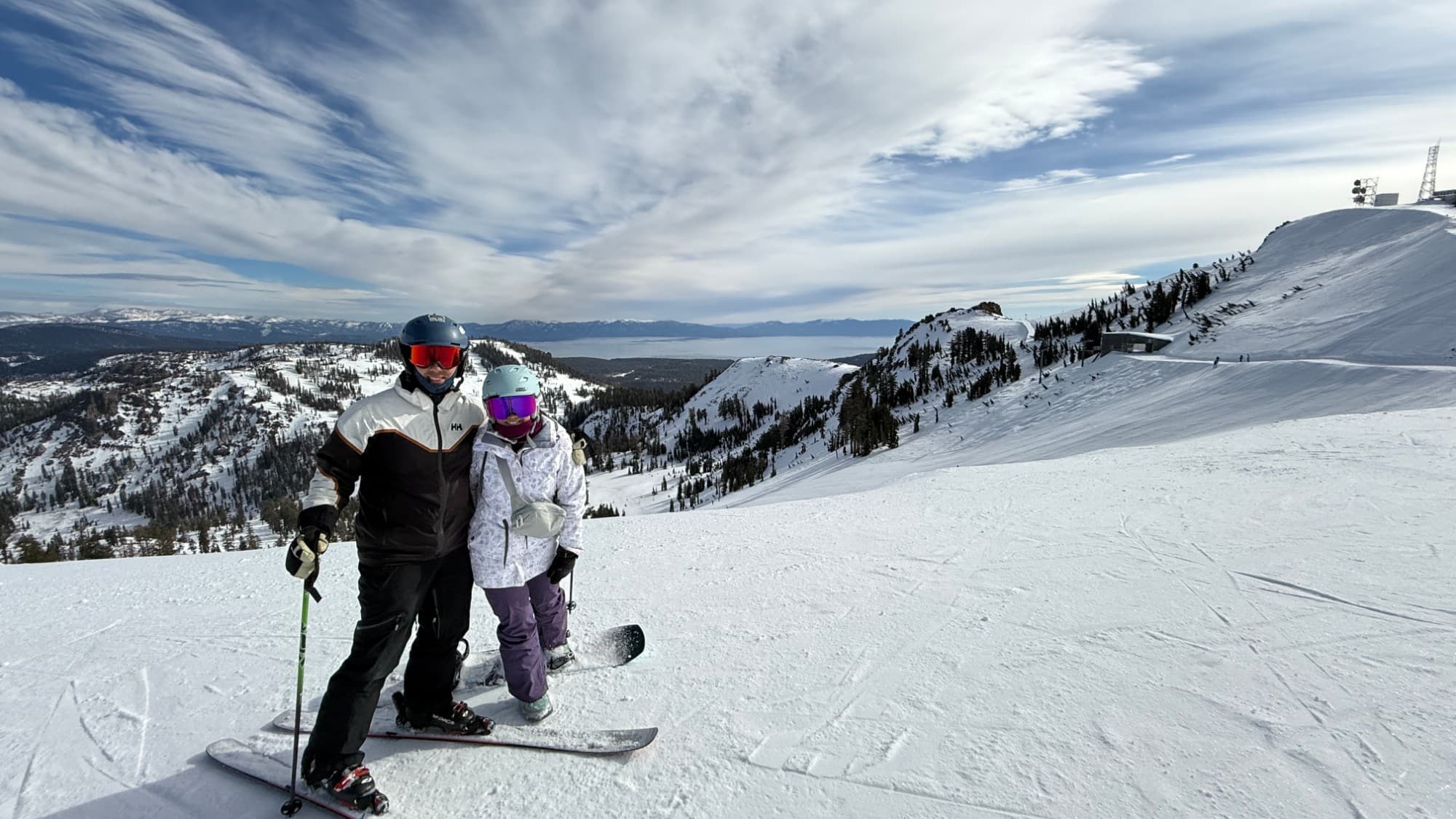 Kevin on skis beside a snowboarder at the top of a run, with snowy ridges and a lake far below